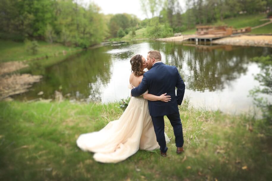 Bride and groom kissing by the lake at Caribou Bay Retreat wedding venue in Central Wisconsin, with greenery and water reflecting the romantic outdoor setting.