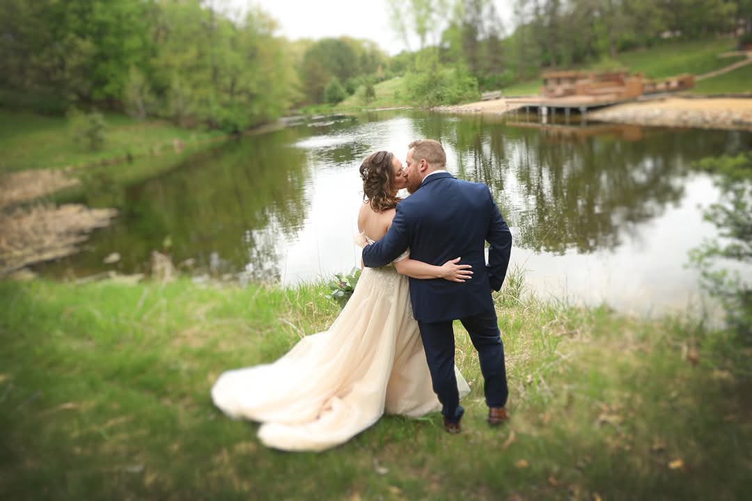 Bride and groom kissing by the lake at Caribou Bay Retreat wedding venue in Central Wisconsin, with greenery and water reflecting the romantic outdoor setting.