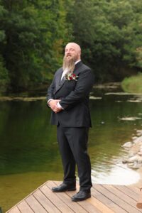 Groom standing on a wooden dock by the lake at Caribou Bay Retreat in a black suit with boutonniere.