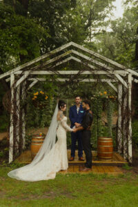 A bride and groom stand hand in hand under a rustic wooden arbor covered in vines, exchanging vows during a summer wedding ceremony at Caribou Bay Retreat in Wisconsin.