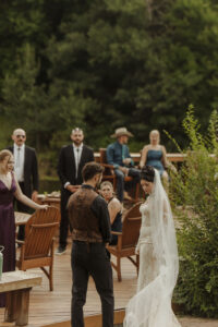 A bride and groom share a quiet moment on a wooden deck surrounded by guests seated in Adirondack chairs during a summer wedding at Caribou Bay Retreat.