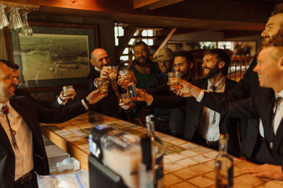 Central Wisconsin wedding venue near Stevens Point at Caribou Bay Retreat – the groom and groomsmen share a toast at the rustic bar before the ceremony, laughing together in a warm wood-beamed lodge setting.