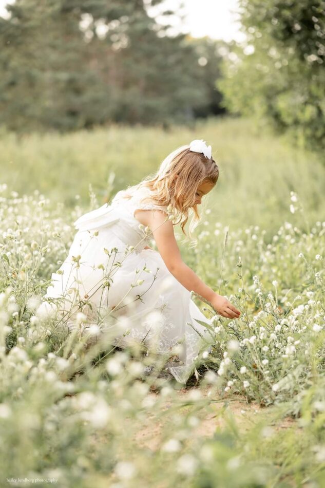 Central Wisconsin wedding venue near Stevens Point at Caribou Bay Retreat – young flower girl in a white dress picking wildflowers in a sunlit meadow.