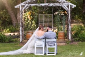 Central Wisconsin wedding venue near Portage at Caribou Bay Retreat – bride and groom seated together facing the outdoor chapel, veil flowing across the grass beneath greenery and wooden barrels.