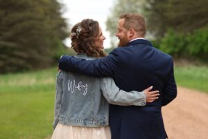 Central Wisconsin wedding venue near Stevens Point at Caribou Bay Retreat – newly married couple walking arm in arm down a wooded path, bride wearing a “wifey” denim jacket after the ceremony