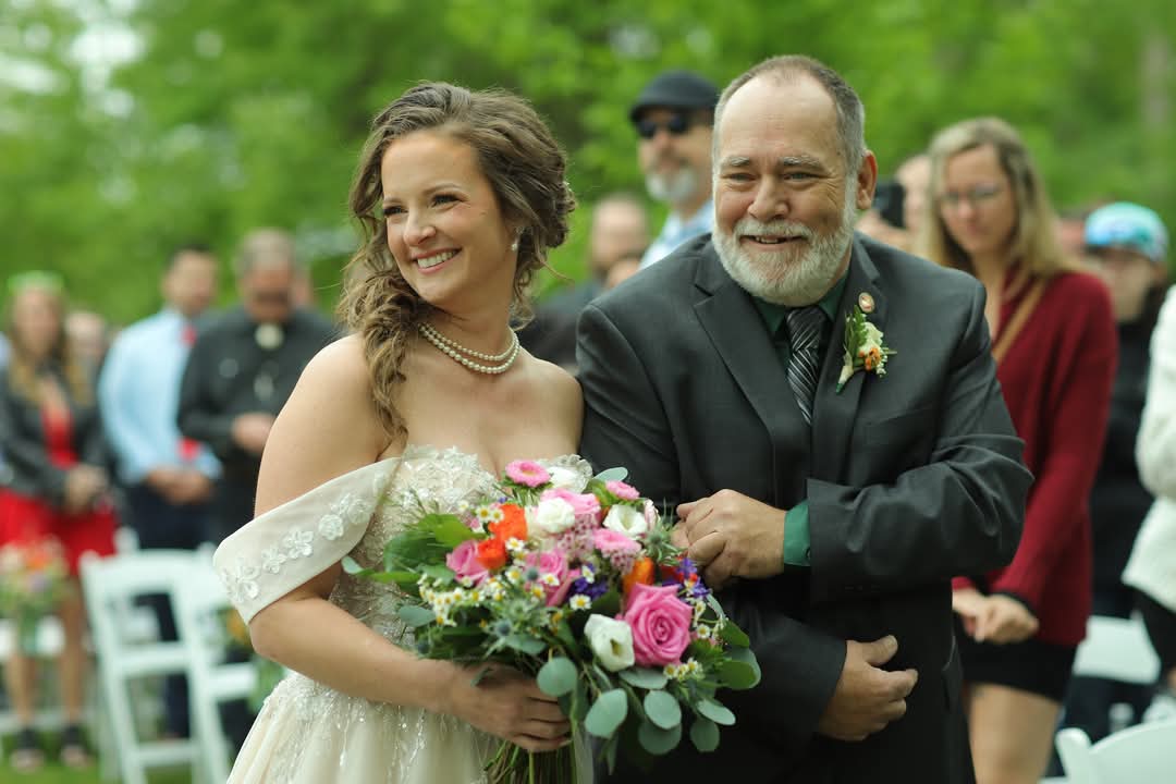 Central Wisconsin wedding venue near Stevens Point at Caribou Bay Retreat – emotional outdoor ceremony moment as a bride walks down the aisle with her father, holding a colorful bouquet surrounded by loved ones