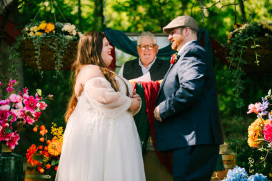 Central Wisconsin wedding venue near Stevens Point at Caribou Bay Retreat – bride and groom smiling and holding hands during an intimate outdoor chapel ceremony surrounded by colorful flowers and woodland greenery.