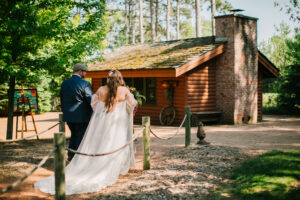 Central Wisconsin wedding venue near Wisconsin Dells at Caribou Bay Retreat – bride and groom walking hand in hand along a wooded path toward the outdoor chapel beside a rustic log cabin.