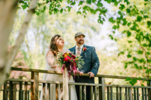 Central Wisconsin wedding venue near Oshkosh at Caribou Bay Retreat – bride and groom standing together on a wooden bridge beneath leafy trees holding a vibrant bouquet.