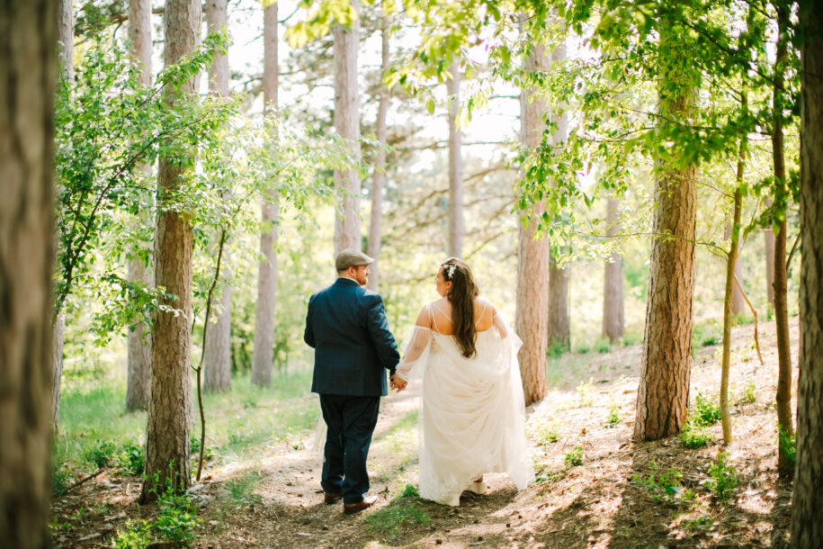 Central Wisconsin wedding venue near Appleton at Caribou Bay Retreat – bride and groom walking hand in hand along a sunlit woodland trail surrounded by tall pine trees.