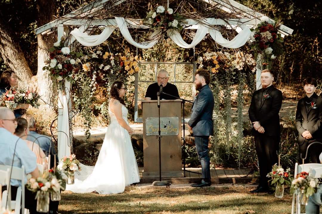 Central Wisconsin wedding venue near Wisconsin Dells at Caribou Bay Retreat – outdoor lakeside ceremony under a rustic arbor with twinkle lights and floral greenery as bride and groom exchange vows